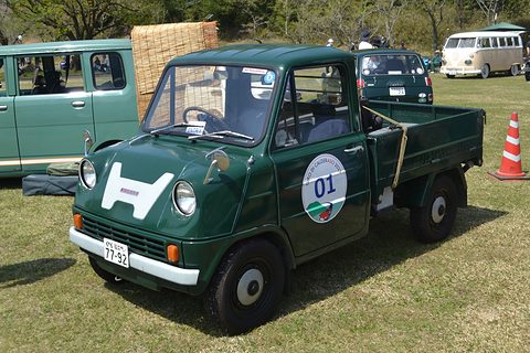 Green pickup on grass with a round 01 sticker on the door, a traffic cone and a van behind. Opens a larger view