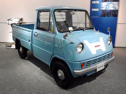 Pale blue pickup in a white gallery, a bare engine on a stand behind its bed and a blue panel of dates.