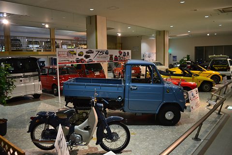 Wide view of a museum hall: a blue tracked pickup, a red and white motorcycle in front, banners above.