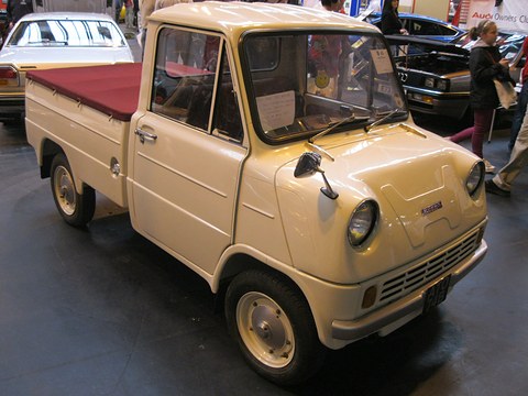 Cream pickup with a red cover over its bed, round headlamps and wing mirrors, on a dark show floor. Opens a larger view