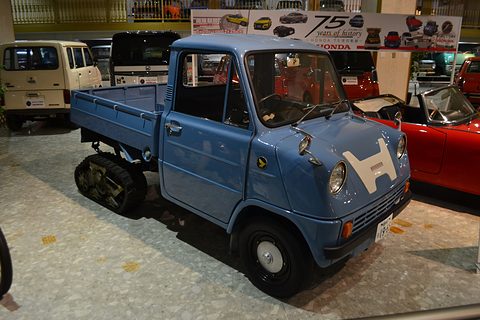 Blue pickup with a white H badge, round headlamps and crawler tracks at the rear, under a Honda anniversary banner. Opens a larger view