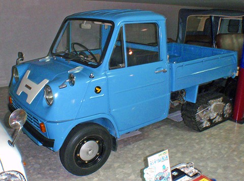 Light blue pickup on a museum floor, its rear wheels replaced by rubber crawler tracks.