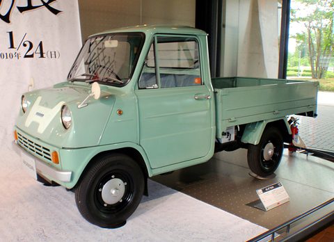 Pale green pickup on a low plinth in a gallery, a large poster with black lettering behind.