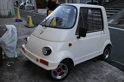 Tiny white single-seat car with round headlamps, a curved windscreen and a door, parked on a forecourt beside traffic cones.