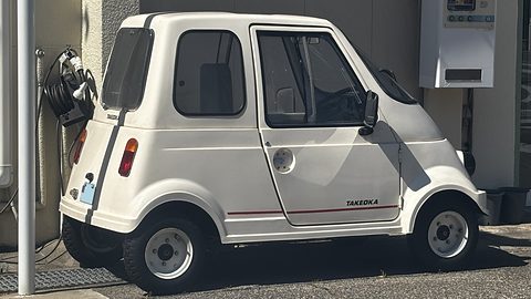 Side view of a white tiny single-seat car with a sliding door and steel wheels by a wall with a meter box.
