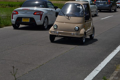 Small beige rounded single-seat car driving along a country road behind a white roadster, rice fields beside the road. Opens a larger view