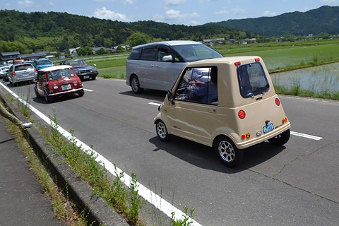Rear of a small beige rounded single-seat car on a country road, a grey van and other cars ahead, hills behind. Opens a larger view