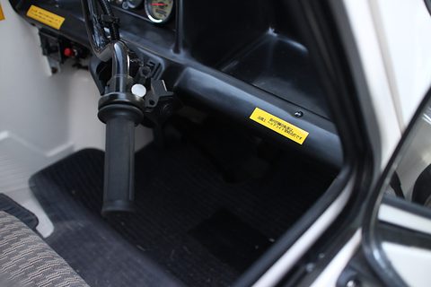 Inside a tiny car: a motorcycle-style twist grip on a column, a black floor mat and a yellow warning sticker. Opens a larger view