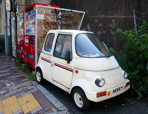 White tiny single-seat car with round headlamps and a red stripe parked on a pavement beside drinks machines. Opens a larger view