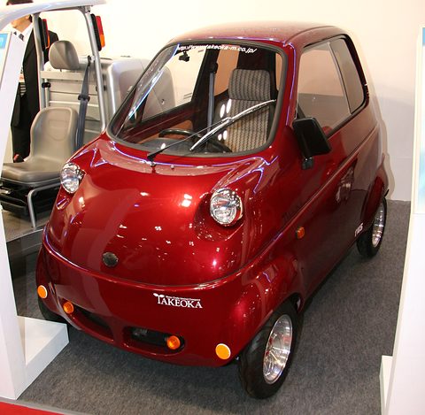 Glossy dark red tiny single-seat car with round headlamps and a Takeoka badge, on a white show stand.