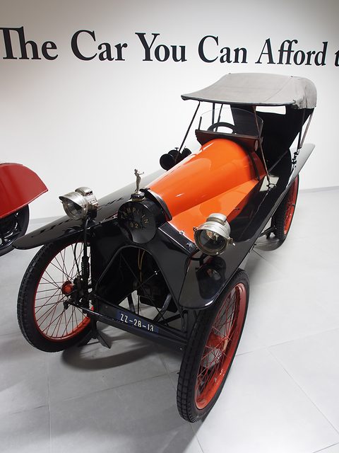 A narrow red and black tandem cyclecar with a grey hood and red wire wheels on a white floor. Opens a larger view