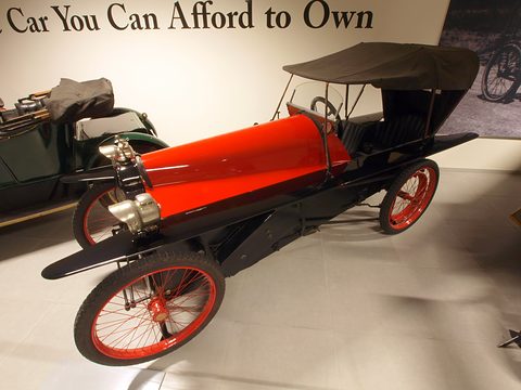 A narrow tandem cyclecar with a red bonnet, black body, folding hood and red wire wheels under a sign reading The Car You Can Afford to Own. Opens a larger view