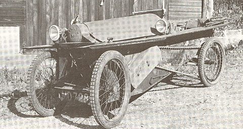 A narrow cyclecar with wire wheels, twin headlamps and a long exposed chain along its side, parked on rough ground.