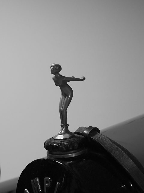 A small bronze statuette of a woman leaning forward with arms outstretched, standing on an ornate radiator cap. Opens a larger view