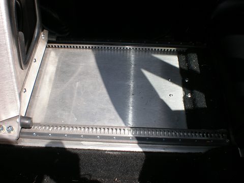 Brushed aluminium floor panel with a toothed seat rail, in a dark car interior with black carpet. Opens a larger view