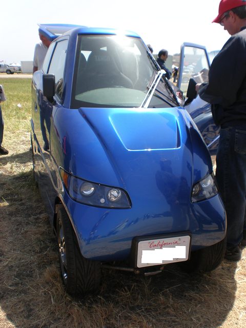 Front of a very narrow blue car with a blanked California plate, a man in a red cap beside its open door. Opens a larger view
