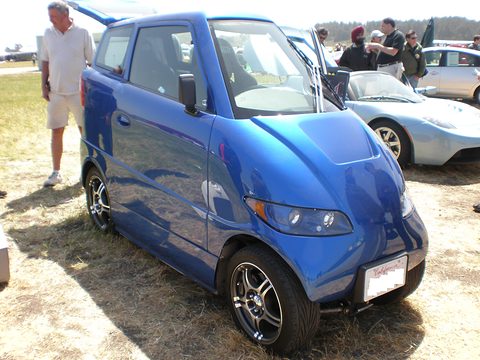 Narrow blue two-seat car with its doors open on dry grass, visitors and other cars behind. Opens a larger view