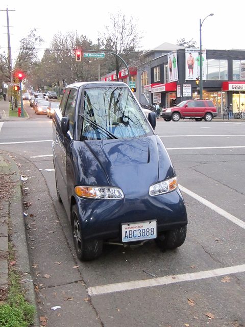 Very narrow dark blue two-seat car parked head-on at a kerb with autumn leaves, shops and traffic lights behind.