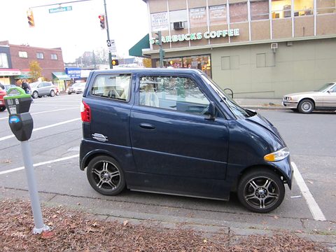 Narrow dark blue two-seat car parked by a parking meter on a corner, a coffee shop behind. Opens a larger view