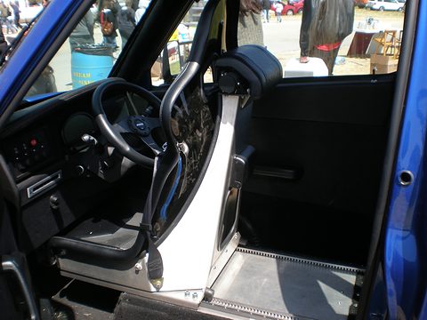 Black racing seat with a tubular roll cage, a steering wheel and a bare aluminium sill seen through the open door.