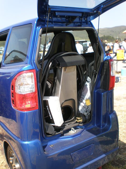 Rear of a narrow blue car with the hatch raised, a seat and a box inside, people at a show behind. Opens a larger view