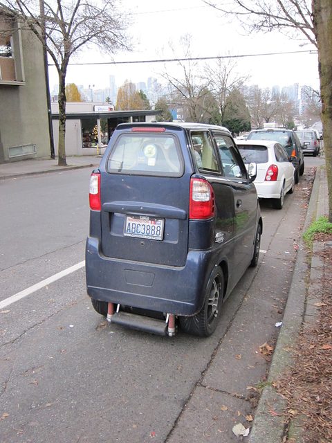 Rear of a tall, very narrow dark blue car with a Washington plate, parked at a kerb behind a white car. Opens a larger view
