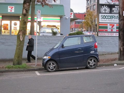 Side view of a short, tall dark blue car at a kerb, a man walking past a convenience store behind it.