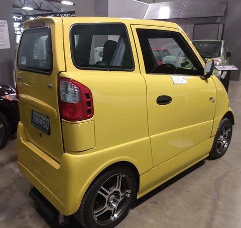 Rear of a narrow yellow two-seat car with tall tail lamps on a museum floor, information panels behind. Opens a larger view