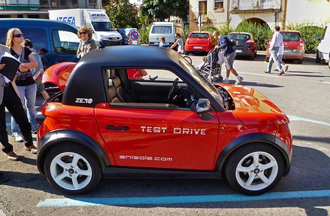 Side and rear of a red two-seat coupé with a black roof and Test Drive lettering, passers-by on the square. Opens a larger view