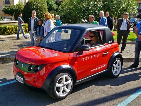 Red two-seat coupé with a black roof and Test Drive lettering, a group of onlookers standing behind it. Opens a larger view
