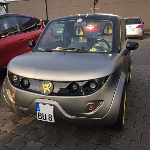 Front of a matt grey two-seat coupé with yellow wheels and a large ZZ badge, parked on dark paving. Opens a larger view