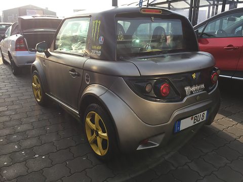 Rear of a matt grey two-seat coupé with yellow wheels and sponsor stickers, parked on dark paving. Opens a larger view
