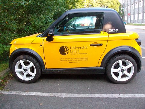 Side view of a yellow two-seat coupé with a black roof and university lettering on its door, parked by trees.