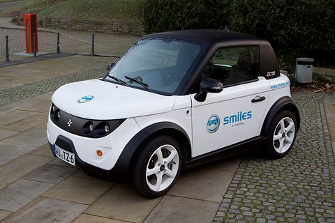 White two-seat coupé with a black roof and blue Smiles lettering, parked on stone paving.