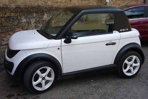 White two-seat coupé with a black roof and alloy wheels parked by an old brick wall. Opens a larger view