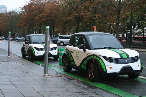 Two white, green and black two-seat coupés plugged into charging posts in green bays beside a tree-lined avenue.