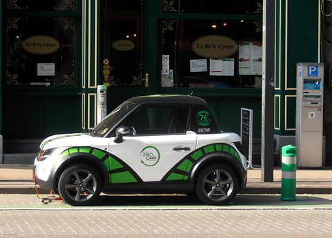 Side view of a white two-seat coupé with green stripes parked in front of a dark green shopfront. Opens a larger view