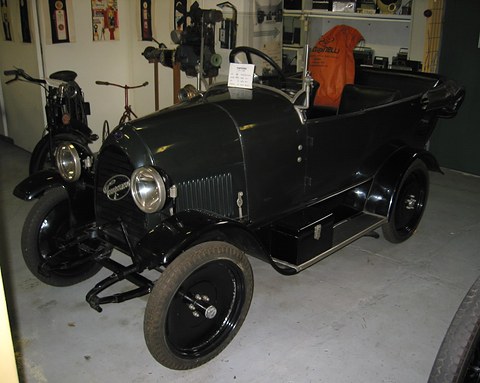 Black open tourer with large headlamps and black disc wheels in a museum of old radios and machines.