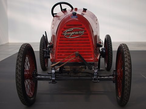 Head-on view of a red cyclecar with a slatted cowl, script badge and wire wheels. Opens a larger view