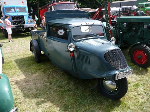 A weathered blue-grey three-wheeler with a mesh grille, single front wheel and a pick-up bed on grass. Opens a larger view