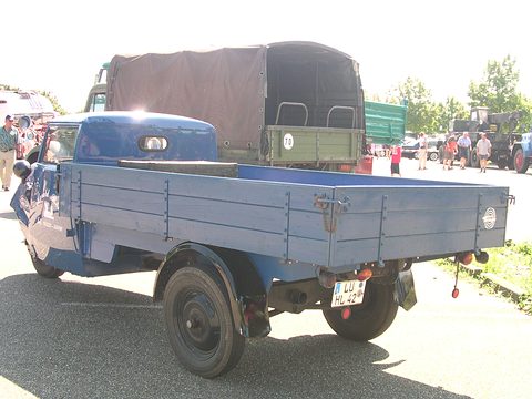 Rear three-quarter view of a long blue drop-side bed on two rear wheels behind a small rounded cab. Opens a larger view