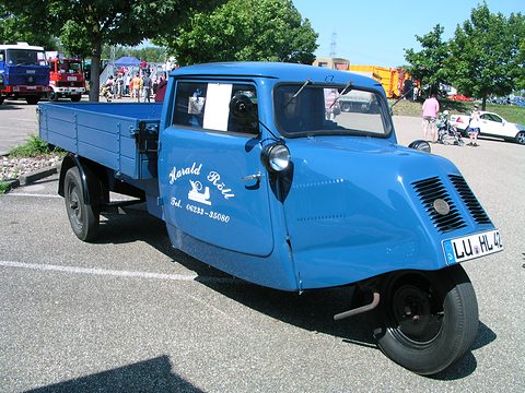 A bright blue three-wheeler with a signwritten door, single front wheel and a long drop-side bed on tarmac. Opens a larger view