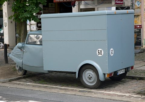Rear three-quarter view of a grey box van with 60 speed discs on its back and side, on a cobbled kerb. Opens a larger view