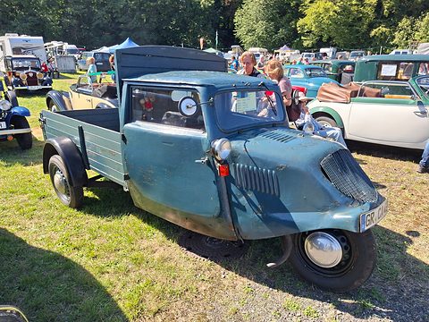 A faded blue-grey three-wheeled pick-up with a rounded nose and a single front wheel on a sunny field. Opens a larger view