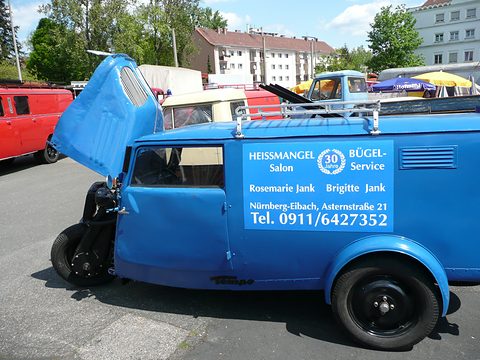 A blue three-wheeled box van with signwritten panels and its rounded nose lifted open, parked in a street. Opens a larger view