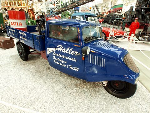 A blue three-wheeler with a rounded cab, signwritten doors and a single front wheel, a drop-side bed behind.