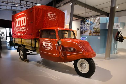 A red three-wheeled van with a curved cab and a canvas-covered box lettered OTTO Versand GmbH in a museum hall. Opens a larger view