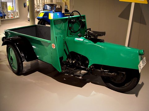 A green open three-wheeled truck with its load bed at the front and the driver’s seat behind it.