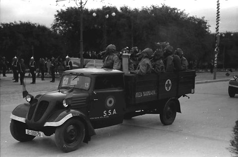 A dark small truck with a curved nose and a Red Cross on its tailboard, men in gas masks seated in the back. Opens a larger view