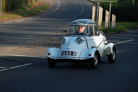 Pale blue bubble car driving towards the camera on a curving main road in low autumn sun, its driver visible under the canopy. Opens a larger view